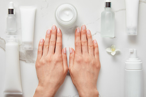 cropped view of woman hands near cosmetic glass bottles, jar with cream, moisturizer tubes, dispenser and jasmine on white surface