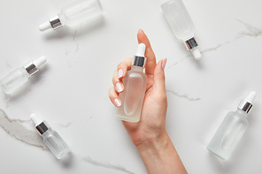 cropped view of woman hand with moisturizer serum near cosmetic glass bottles on white surface