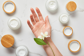 cropped view of jasmine flowers on woman hand near jars with cosmetic cream on white surface