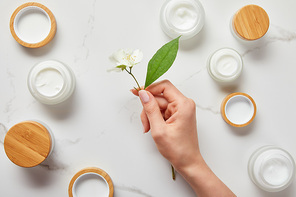 cropped view of woman hand with jasmine flowers over jars with cosmetic cream on white surface