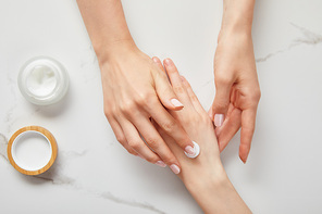 cropped view of woman applying cream on hand, using white moisturizer from jar on white surface