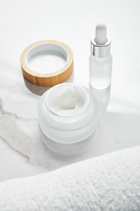 opened jar with cosmetic cream and glass bottle near towel on white surface