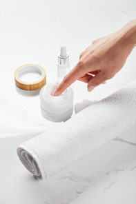 cropped view of woman using cosmetic cream on white surface with glass bottle and towel