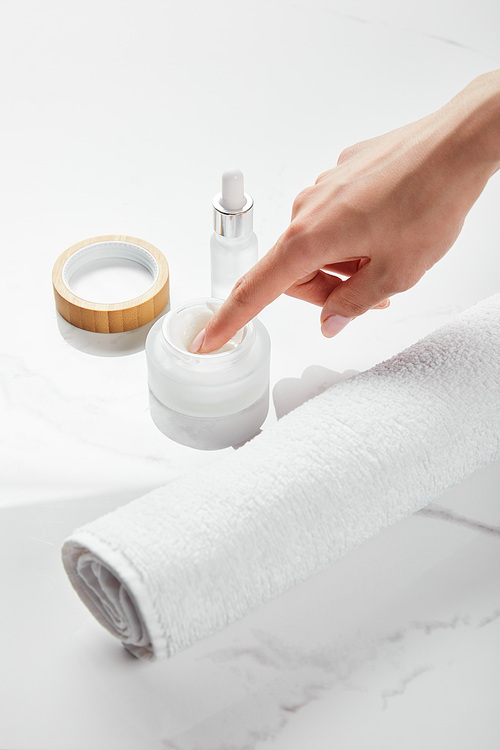 cropped view of woman using cosmetic cream on white surface with glass bottle and towel