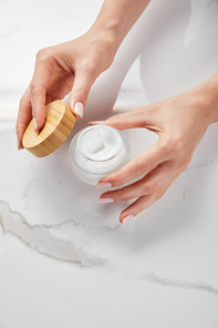 cropped view of woman opening jar with cream on white surface