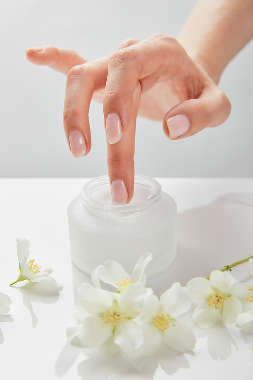 cropped view of woman hand touching cream in jar near jasmine flowers on white surface