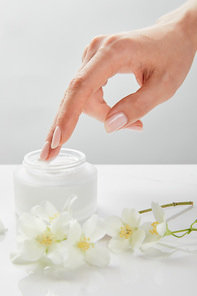 cropped view of woman hand touching cream in jar near jasmine flowers on white surface