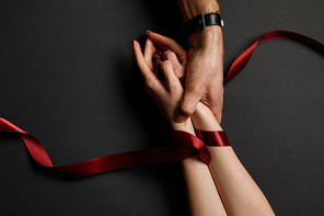 cropped view of man holding female hands in red satin ribbon on black background