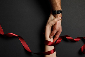 cropped view of man and woman in red satin ribbon holding hands on black background