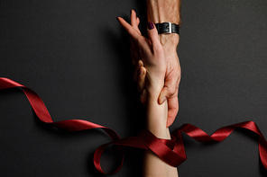 cropped view of man holding female hand in red satin ribbon on black background