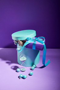 blue box Christmas decoration and present near hourglass on purple background