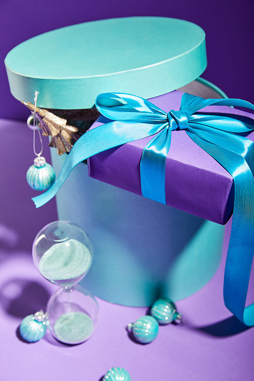 selective focus of blue box Christmas decoration and present near hourglass on purple background