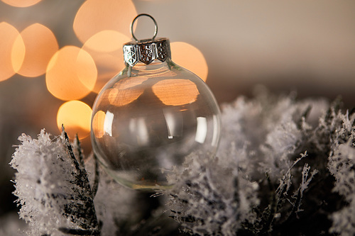 decorative transparent christmas ball on spruce branches in snow with blurred yellow lights