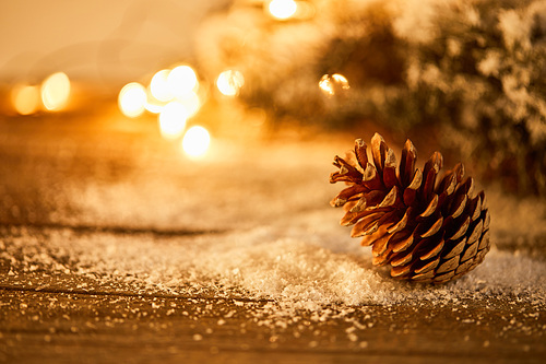 decorative pine cone on wooden table with spruce branches in snow and christmas lights bokeh