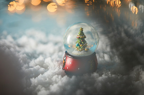 selective focus of decorative snowball with christmas tree standing on blue in snow with lights bokeh
