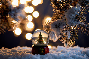 decorative christmas tree in snowball standing in snow with spruce branches and blurred lights at night