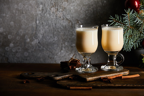 glasses with delicious eggnog drink on cutting board near spruce branch on grey stone background