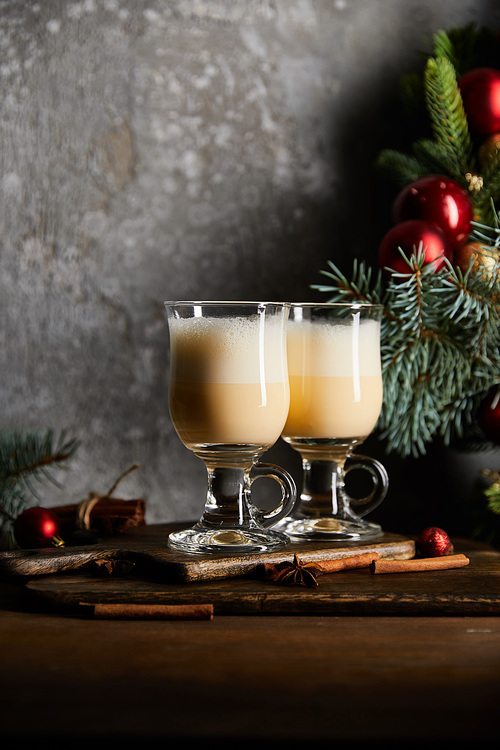 glasses with delicious eggnog drink on cutting board near decorated spruce branch on grey stone background