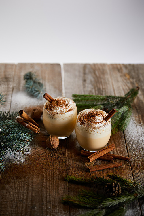 eggnog cocktail with whipped cream and cinnamon near spruce branches and powdered sugar scattered on wooden table isolated on grey