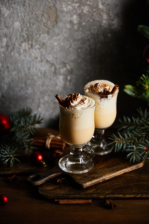 delicious eggnog cocktail with whipped cream and cinnamon on cutting board near spruce branches on grey stone background