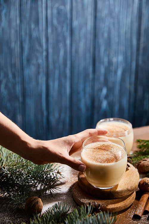 cropped view of woman taking glass with eggnog cocktail near spruce branches on blue wooden background