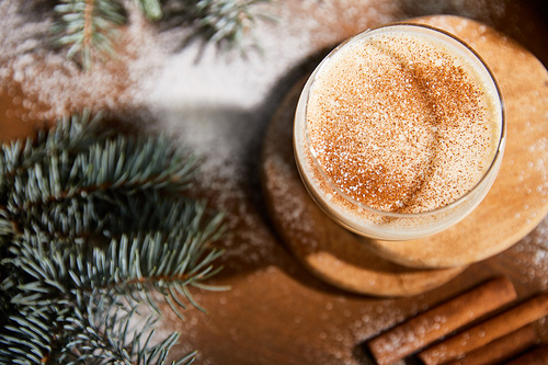 top view of delicious eggnog cocktail on round wooden board near spruce branches and scattered sugar powder