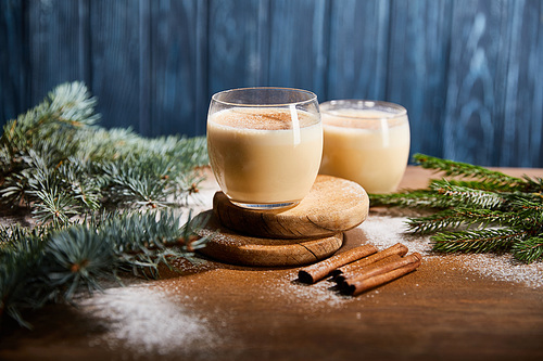 delicious eggnog cocktail on round wooden boards near spruce branches, cinnamon sticks and scattered sugar powder on blue textured background