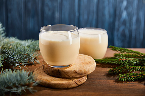 delicious eggnog cocktail on round wooden boards near spruce branches on blue textured background