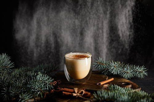 tasty eggnog cocktail on cutting board near spruce branches and cinnamon sticks on black background with powdered sugar falling like snow