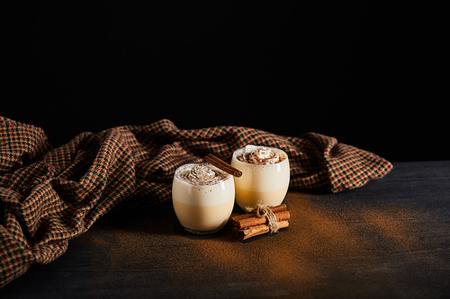 delicious eggnog cocktail, cinnamon sticks and checkered cloth on table covered with cinnamon powder isolated on black
