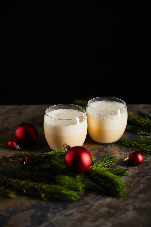traditional eggnog cocktail near spruce branch and Christmas balls on dark marble surface isolated on black