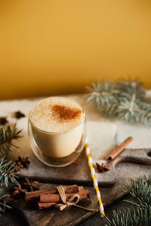 traditional eggnog cocktail on chopping board covered with sugar powder, spruce branches, cinnamon sticks and straw on orange background