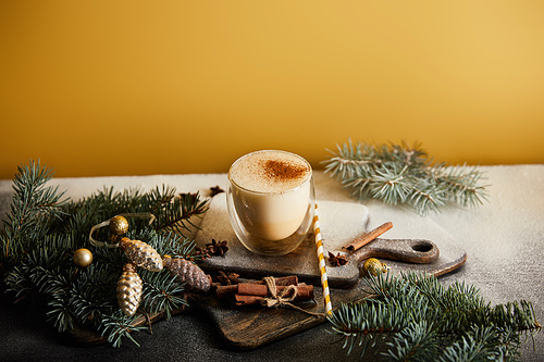 eggnog cocktail on chopping board covered with sugar powder, spruce branches, cinnamon sticks, straw and Christmas baubles on orange background