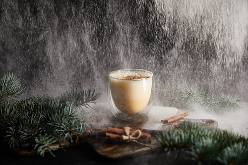 tasty eggnog cocktail on cutting board near spruce branches and cinnamon sticks on black background with powdered sugar falling like snow