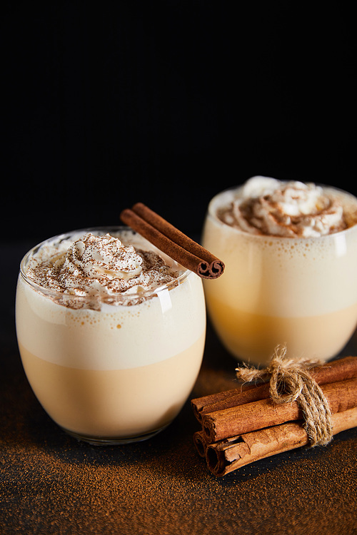 delicious eggnog cocktail with whipped cream and cinnamon sticks on table covered with cinnamon powder isolated on black