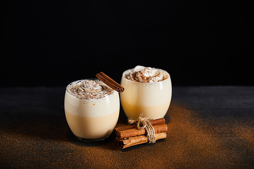 traditional eggnog cocktail with whipped cream and cinnamon sticks on table covered with cinnamon powder isolated on black