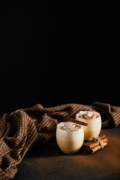eggnog cocktail with whipped cream, cinnamon sticks and checkered cloth on table covered with cinnamon powder isolated on black