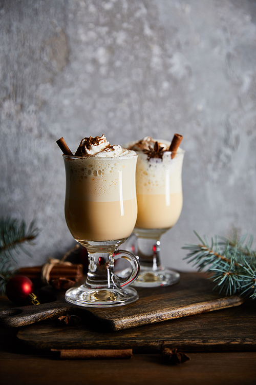 traditional eggnog cocktail with whipped cream and cinnamon on cutting board near spruce branches on grey stone background