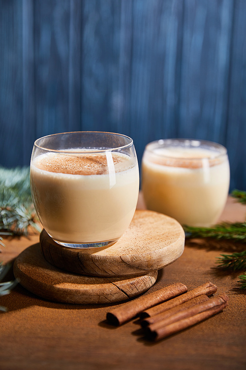 delicious eggnog cocktail on round wooden boards near spruce branches and cinnamon sticks on blue textured background