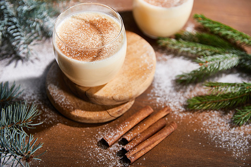 flavored eggnog cocktail on round boards near spruce branches and cinnamon sticks on wooden table covered with sugar powder