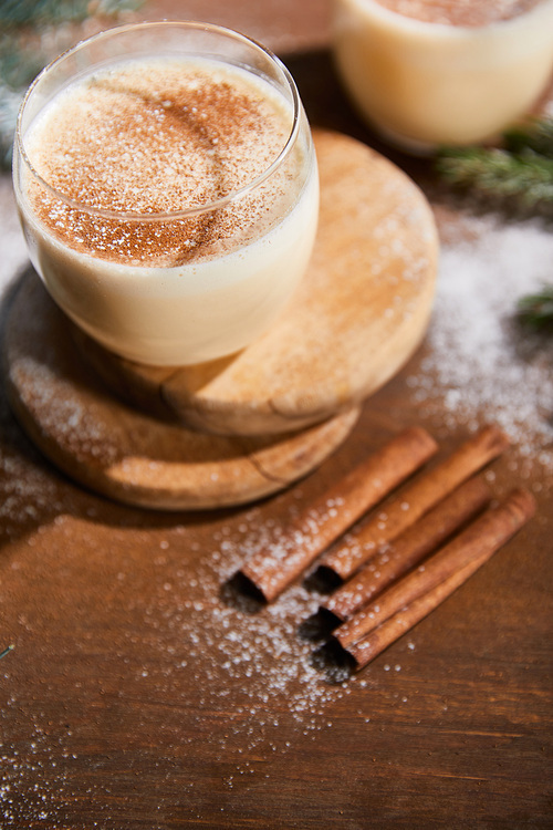 flavored eggnog cocktail on round boards near spruce branches and cinnamon sticks on wooden table covered with sugar powder