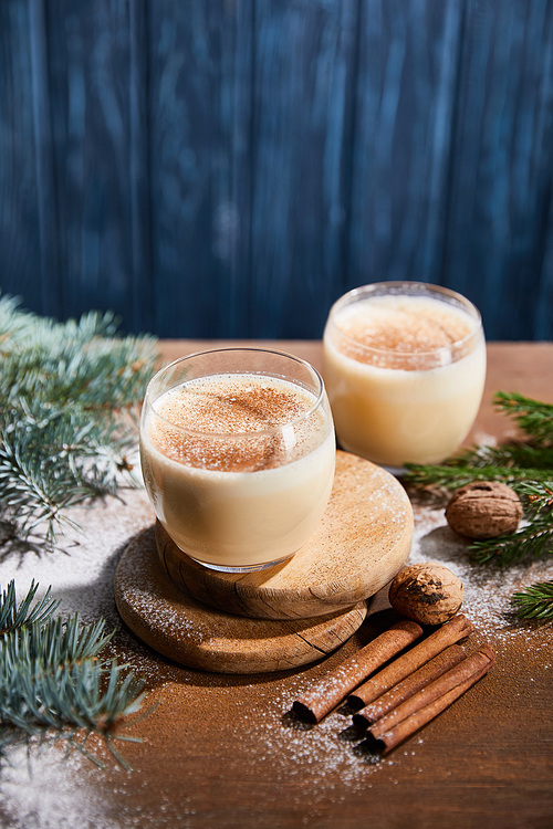 delicious eggnog cocktail, spruce branches, cinnamon sticks and walnuts on wooden table covered with sugar powder on blue textured background