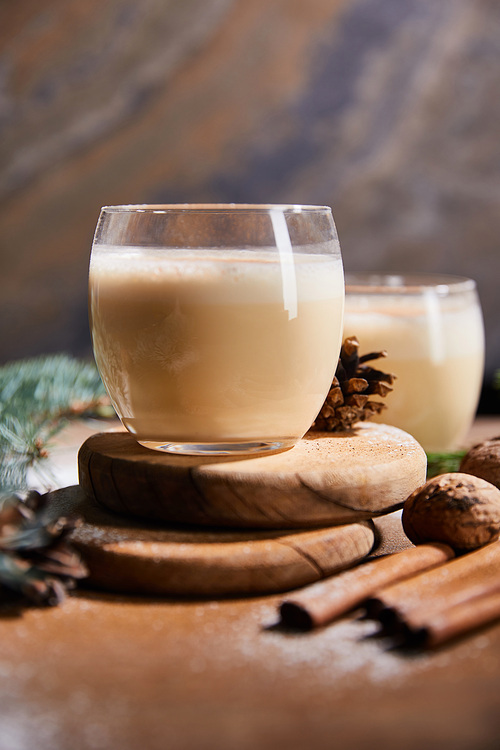 selective focus of delicious eggnog cocktail on round boards, spruce cones, cinnamon sticks and walnuts on marble stone background