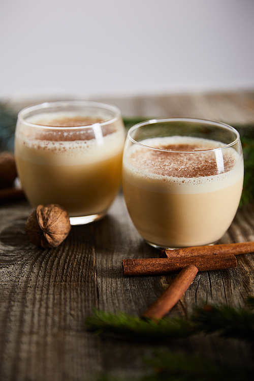 selective focus of flavored eggnog cocktail near cinnamon sticks and walnuts on wooden table isolated on grey