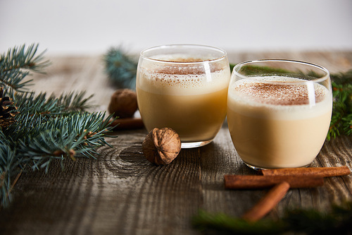 selective focus of flavored eggnog cocktail near cinnamon sticks, walnuts and spruce branches on wooden table isolated on grey