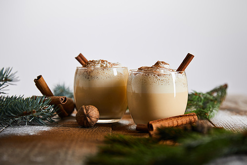 selective focus of eggnog cocktail with whipped cream near cinnamon sticks and spruce branches on wooden table isolated on grey