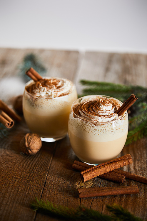 delicious eggnog cocktail with whipped cream near cinnamon sticks and walnuts on wooden table isolated on grey
