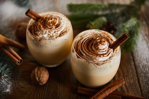 traditional eggnog cocktail with whipped cream near cinnamon sticks, spruce branches and walnuts on wooden table