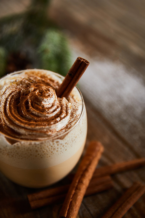 close up view of flavored eggnog drink with whipped cream on wooden table near cinnamon sticks