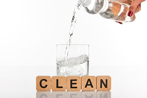 cropped view of woman pouring water from bottle in glass near cubes with clean lettering isolated on white